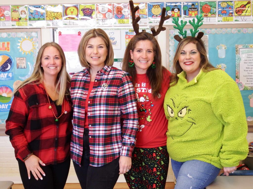 Four women standing wearing holiday apparel in front of a colorful school bulletin board.