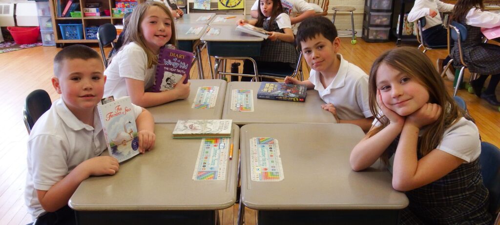 Two boys and two girls in school uniforms sitting at school desks, holding books and smiling at camera