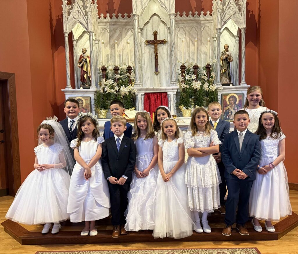 Group of girls and boys in First Communion dresses and suits in front of an altar.